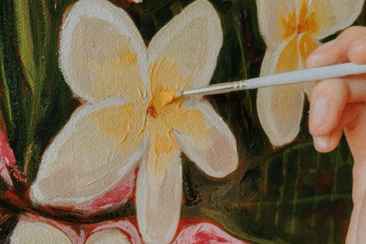 Close-up of a hand holding a fine paintbrush, adding detail to a painted floral scene with white plumeria-like flowers, yellow centers, and dark green leaves.